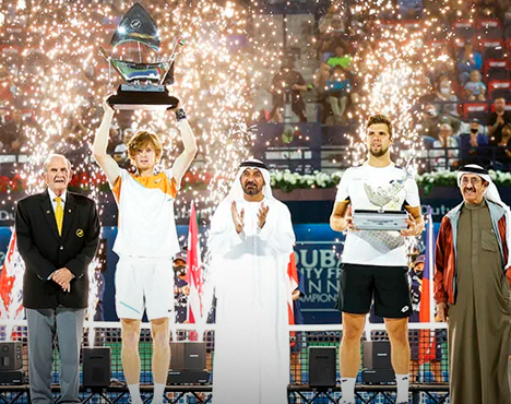 На изображении теннисист с поднятым над головой трофеем, рядом судьи и участники, позади фейерверки. Торжественная награда на спортивном мероприятии