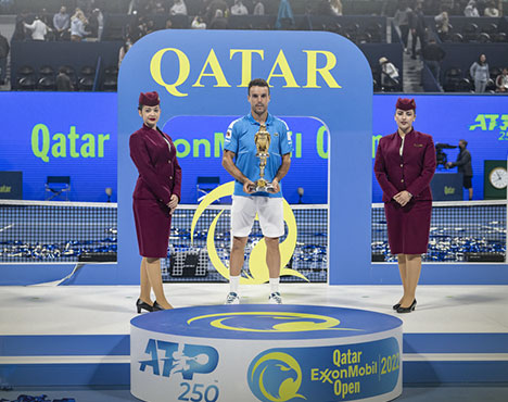 Спортсмен с трофеем стоит на подиуме турнира Qatar ExxonMobil Open. Его окружают две девушки в форме, на заднем плане виден корт и элементы оформления мероприятия